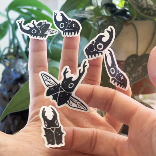 Six individual beetle stickers (removed from the sticker sheet) shown on the tips of fingers against a background with houseplants.