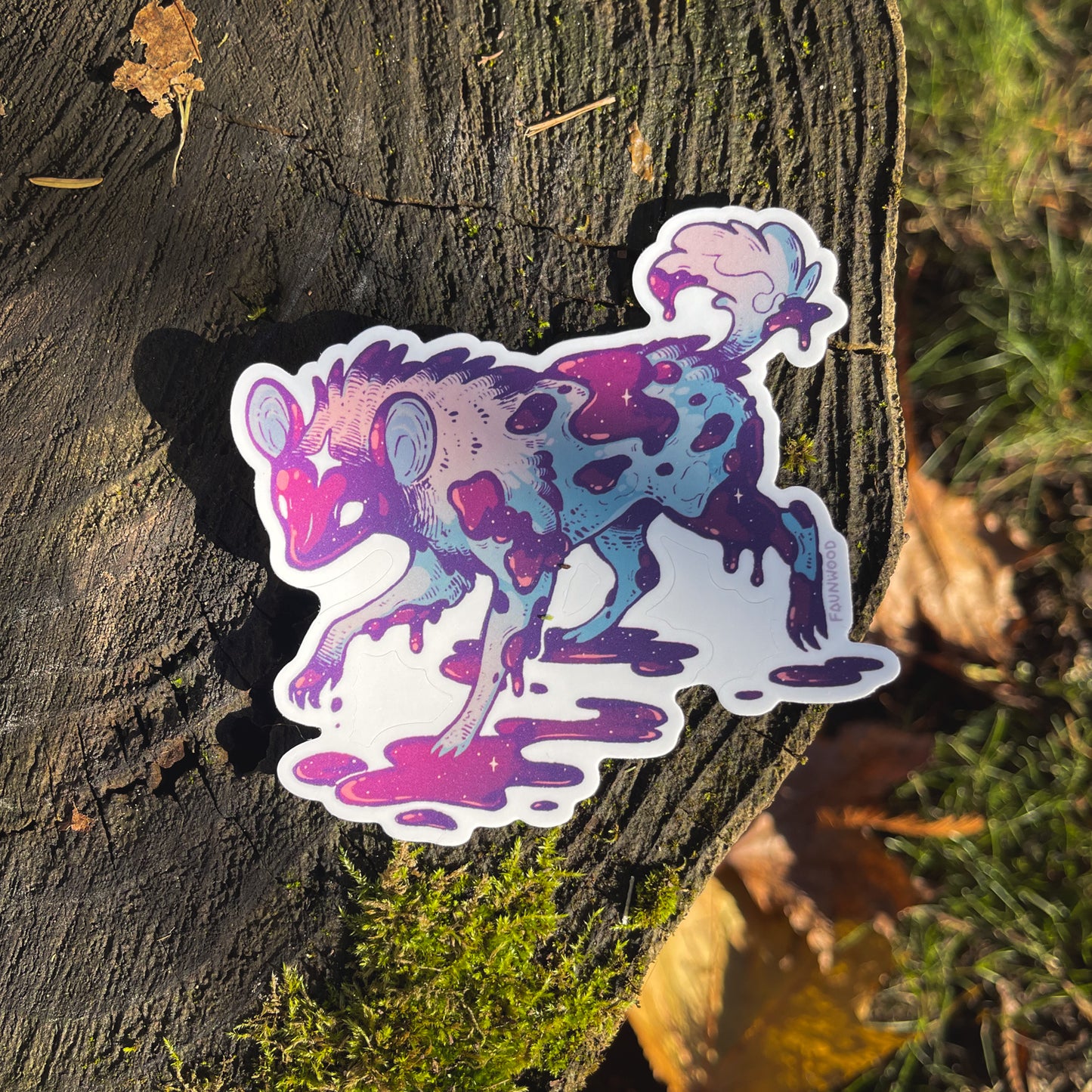A die cut matte vinyl sticker featuring an illustration of an African wild dog with purple and pink coloration, shown in a stylized and dark fantasy-inspired pose, placed on a wooden surface with leaves.