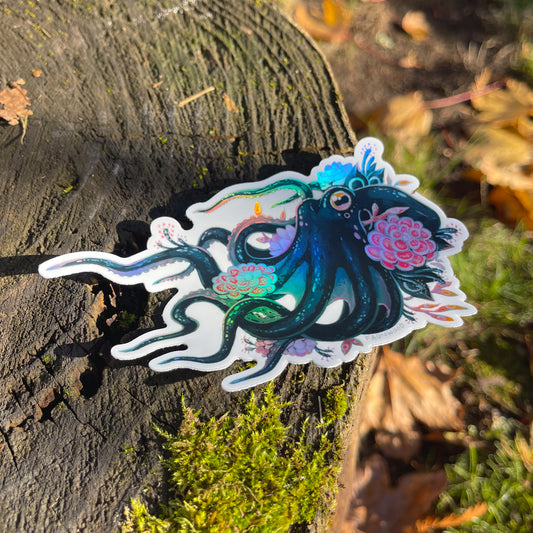 A die cut holographic vinyl sticker depicting a colorful octopus adorned with carnivorous plants. The sticker is photographed on a mossy tree stump.