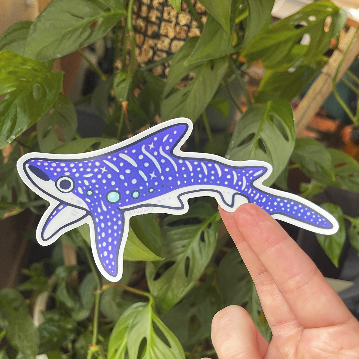 A holo vinyl sticker of a purple illustrated whale shark. The spot and star markings on the whale shark are holographic. In this photo, a hand is holding the sticker in front of a houseplant backdrop.