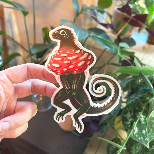 Matte vinyl sticker of a green "frilled lizard" with a red and white mushroom cap in place of frills.  In this photo, a hand is holding the sticker up against a background with houseplants.  The photo is meant to show the space around the tail that appears when you remove the sticker from the backing.