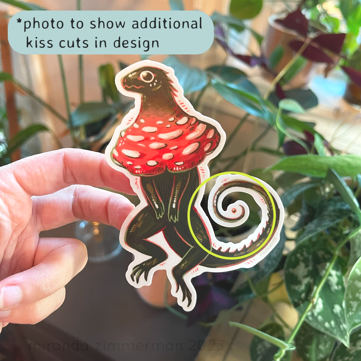 Matte vinyl sticker of a green "frilled lizard" with a red and white mushroom cap in place of frills.  In this photo, a hand is holding the sticker up against a background with houseplants.  The photo is meant to show the space around the tail that appears when you remove the sticker from the backing.