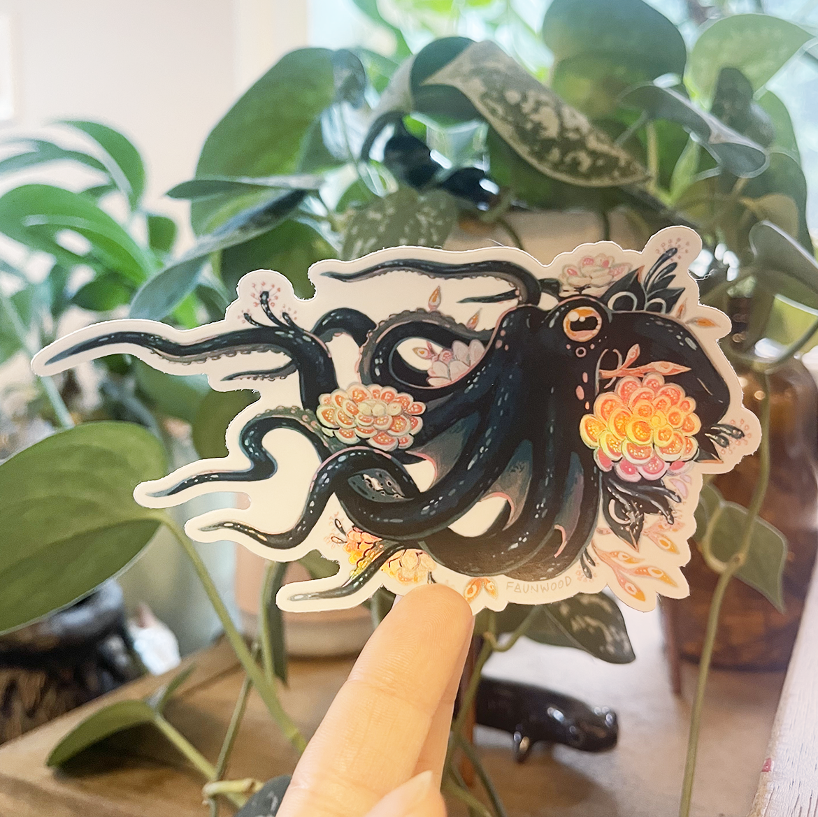 A die cut holographic vinyl sticker depicting a colorful octopus adorned with carnivorous plants.  The sticker is being held by a hand up against a potted plant backdrop.