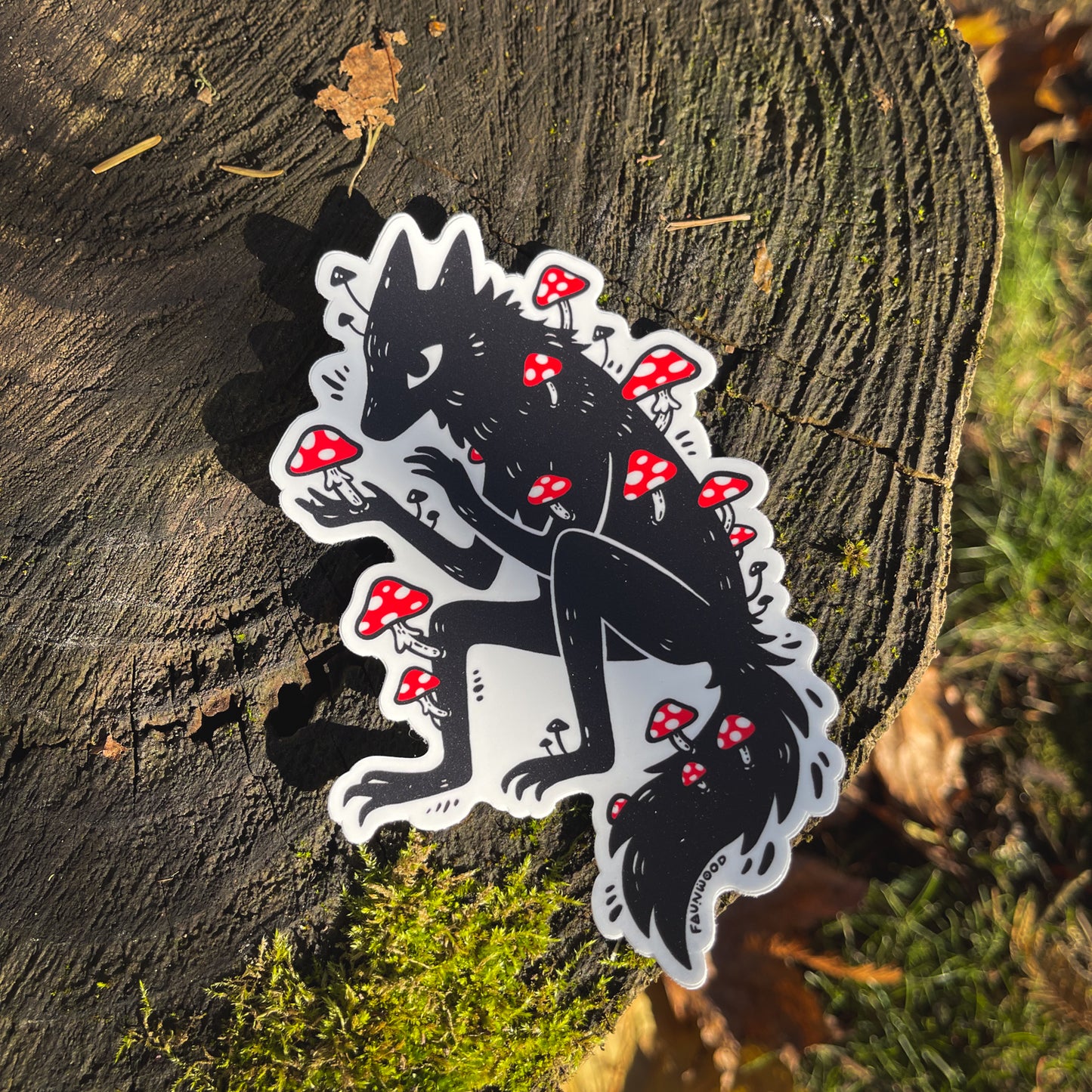 A matte vinyl sticker featuring a black illustration of a wolfy fox covered in red amanita mushrooms.  The sticker is photographed on a mossy tree stump.