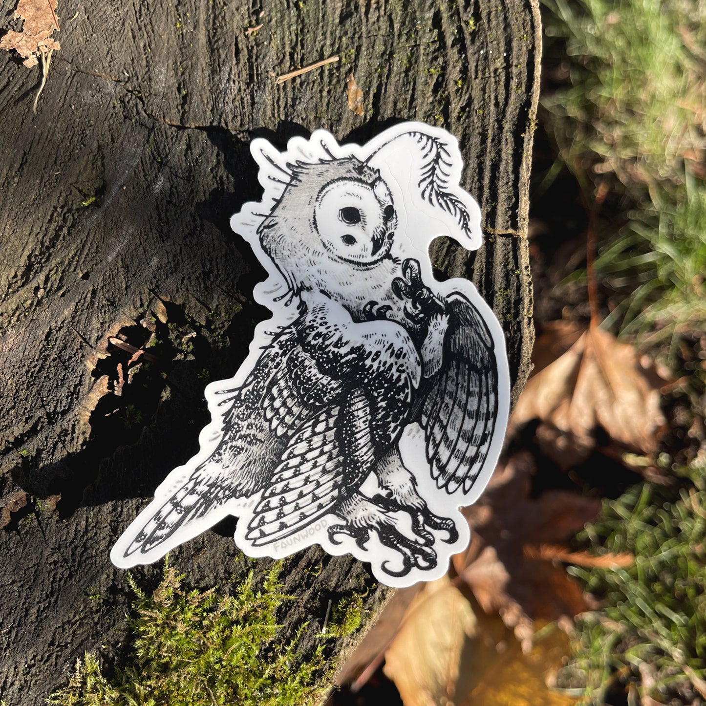 A die-cut matte vinyl sticker of an owl-like fantasy creature, with a black and white color scheme. In this photo, the sticker is resting on a mossy surface with fallen leaves around.