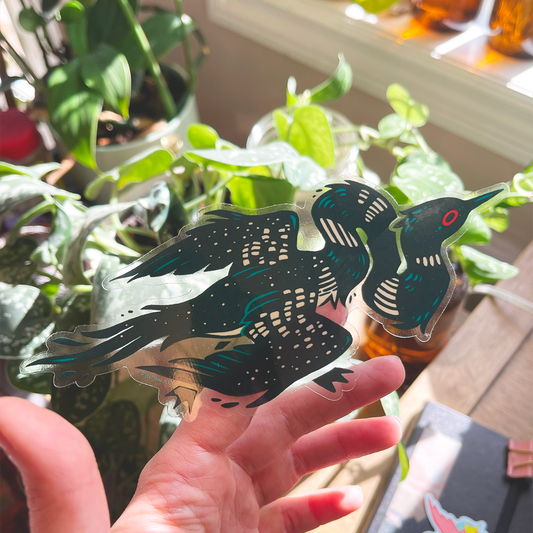 A transparent vinyl sticker of an illustrated Loon with an extra long and twisty neck.  In this photo, a hand is holding the sticker with houseplants in the background, demonstrating the transparent outline.