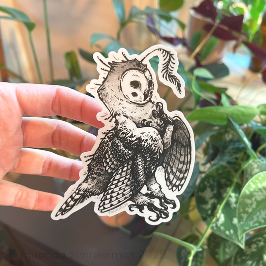 Hand holding a vinyl sticker depicting a black and white owl-creature. 