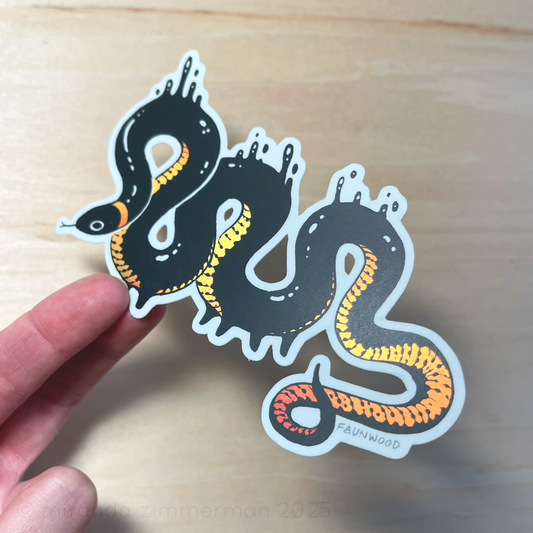 A semi-holo vinyl sticker of an illustrated ringneck snake creature with droplets of black goop.  The snake is black with a yellow-red gradient reflective belly.  In this photo, a hand is holding the sticker under light to show the reflective qualities of the markings.