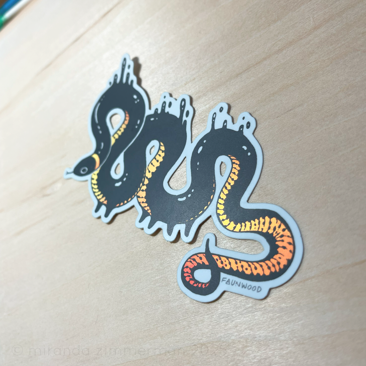 A semi-holo vinyl sticker of an illustrated ringneck snake creature with droplets of black goop.  The snake is black with a yellow-red gradient reflective belly.  In this photo, the sticker is placed under a light to show the reflective qualities of the markings.