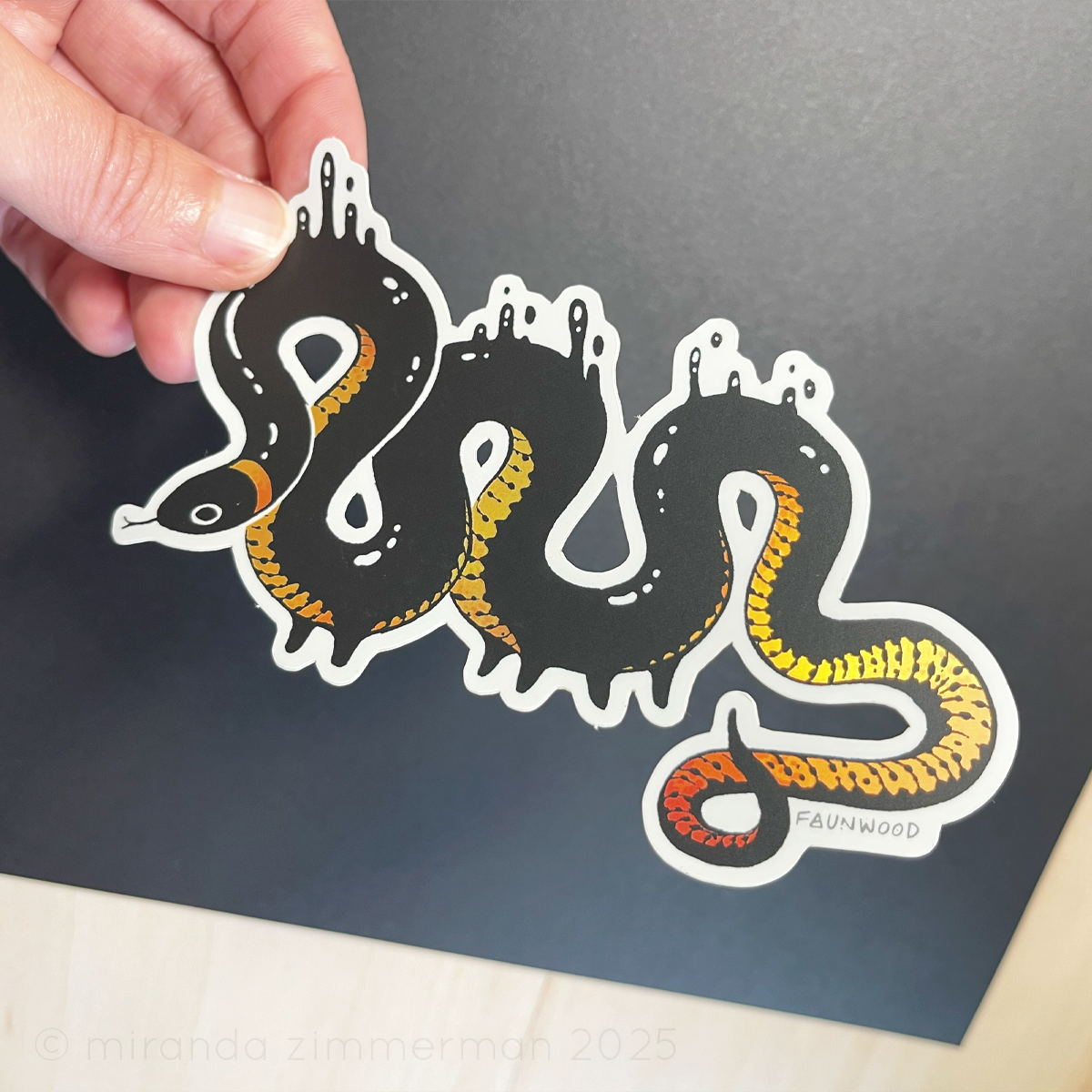 A semi-holo vinyl sticker of an illustrated ringneck snake creature with droplets of black goop.  The snake is black with a yellow-red gradient reflective belly.  In this photo, a hand is holding the sticker under light to show the reflective qualities of the markings.