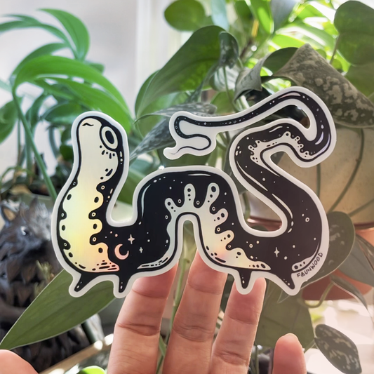 A die-cut holo vinyl sticker of a black and white salamander with star patterns, held by a hand against a backdrop of plants.