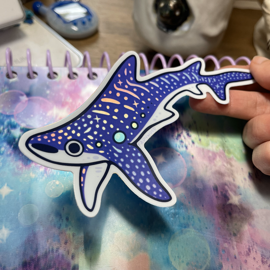 A holo vinyl sticker of a purple illustrated whale shark.  The spot and star markings on the whale shark are holographic.  In this photo, a hand is holding the sticker with a notebook in the background, showing the holographic markings under a light.