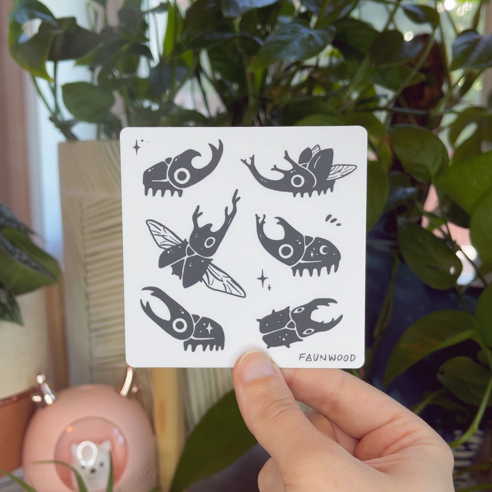 A matte vinyl sticker sheet with six different black beetle illustration stickers.  In this photo, a hand is holding the sheet in front of a background with houseplants.