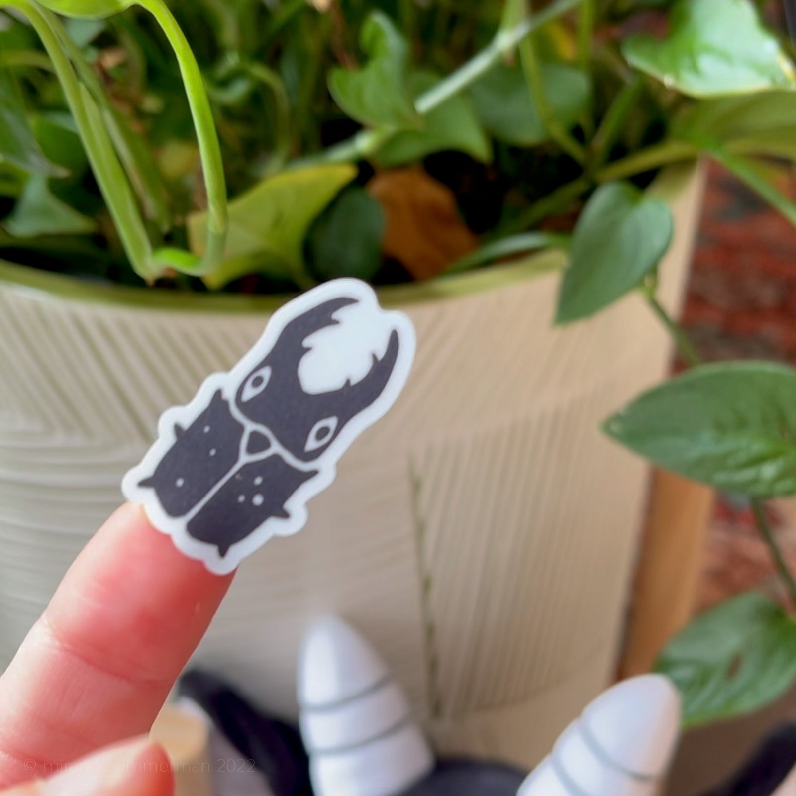 A small individual beetle sticker (removed from the sticker sheet) held by a hand against a blurry houseplant background.